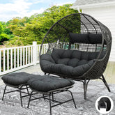 The Morsale Winsford Patio Lounge Chair in black PE wicker, featuring round cushions and crafted with weather-resistant fabric, sits on a deck amidst greenery. Two matching footrests rest on a patterned outdoor rug. A white railing and part of the house are visible.