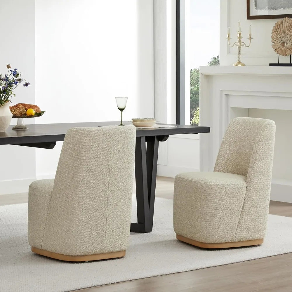 Two Boucle Dining Chairs with Casters by Morsale, embodying a modern home aesthetic, sit at a wooden table with a black base. On the table are a green glass, a bowl, and fruit. A white fireplace with decor is in the background beside a window.