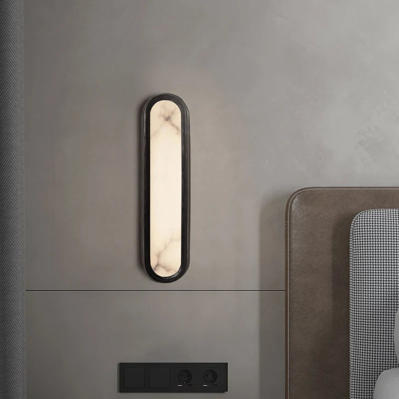 A modern, oval-shaped Moonshade Natural Marble LED Wall Lamp by Morsale with a black frame and soft white light is mounted on a gray wall above a bed with a gray headboard. Below the sconce, there are three black electrical outlets.