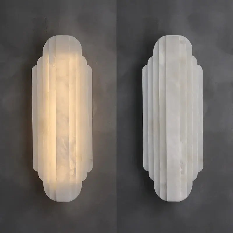 
                  
                    Two white Arina Marble Sconces by Morsale with scalloped edges add sculptural lighting to a dark grey wall; the left sconce is lit, while the right one remains off.
                  
                