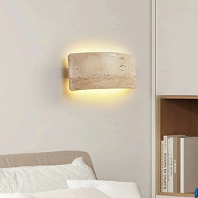 The Natural Travertine LED Wall Light Fixture by Morsale.com is an interior wall sconce featuring a rectangular, curved stone-like cover that emits a warm, soft glow. This energy-efficient LED lighting creates a halo effect on the white wall, emphasizing the texture and natural imperfections of the stone surface.