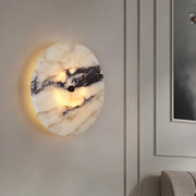 The Moonshade Natural Marble Sconce Light by Morsale, featuring a round marble design with black and white veins, is mounted on a light gray paneled wall. The LED emits a warm glow, illuminating part of a couch and an adjacent framed artwork.