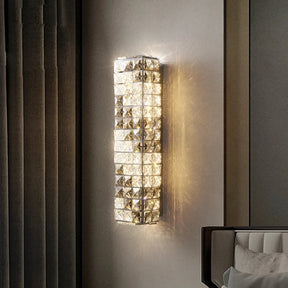 A modern, vertical wall-mounted light fixture with a geometric crystal pattern and integrated LED emits warm light next to a bed. The Bacci Smoke & Clear Crystal Wall Sconce by Morsale is elegantly positioned on the wall. Below the light, there is a bedside table with a framed photo and a small cylindrical object. The bed has pillows and a neatly arranged blanket.