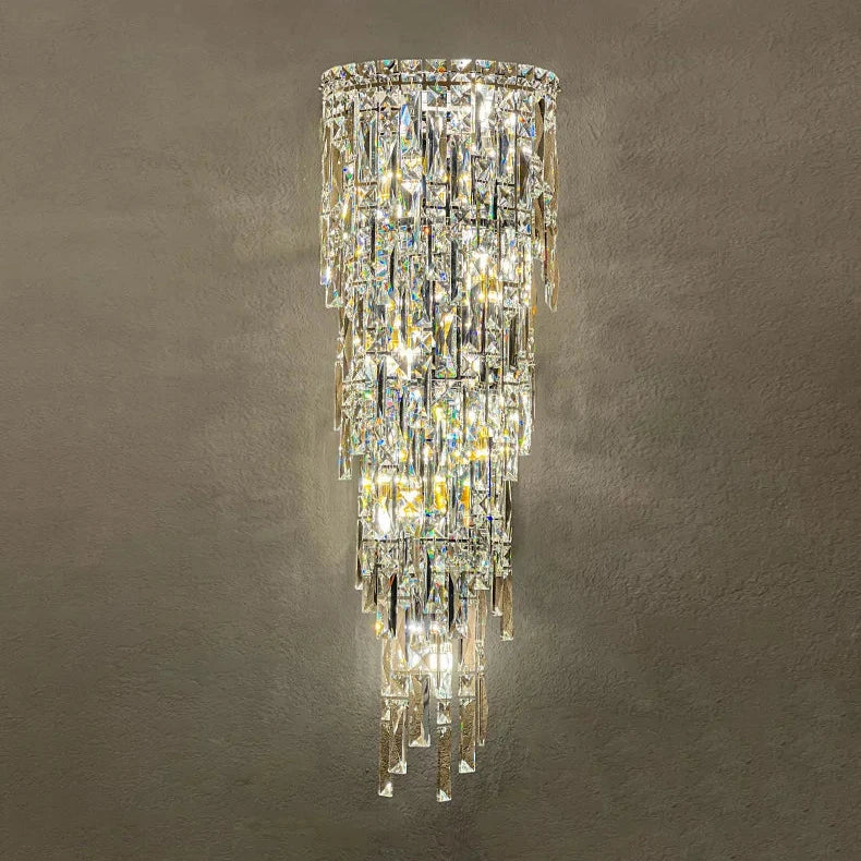 The Bacci Crystal Contemporary Wall Sconce by Morsale is an opulent fixture showcasing handmade crystals gracefully draping against a plain wall. With tiers of cascading crystals and a stainless steel body, it radiates a warm glow that enhances any space with elegance and sophistication.