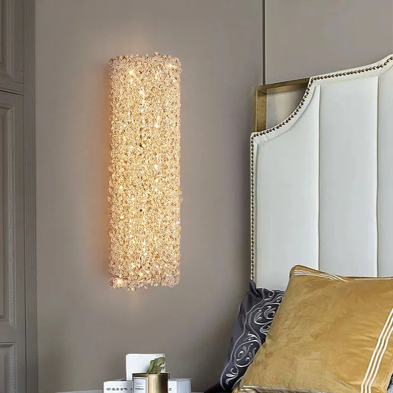 A modern bedroom features the Morsale Bergamo Crystal Sconce, a tall, rectangular wall lamp with a textured design that emits a warm glow next to a white upholstered headboard. A golden pillow and a small plant adorn the bedside table, enhancing the room's contemporary style.