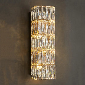 The Bacci Crystal Wall Sconce by Morsale is a modern vertical wall fixture adorned with multiple rectangular crystal prisms, illuminated by LED bulbs. It emits a warm and soft light that reflects beautifully off the crystals, making it stand out when mounted on a dark wall for contrast.