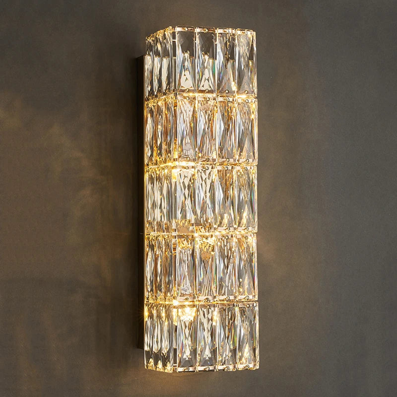 The Bacci Crystal Wall Sconce by Morsale is a modern vertical wall fixture adorned with multiple rectangular crystal prisms, illuminated by LED bulbs. It emits a warm and soft light that reflects beautifully off the crystals, making it stand out when mounted on a dark wall for contrast.