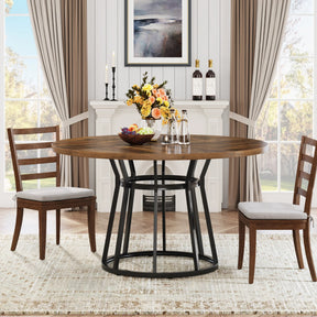 The dining room features a modern style with a Bradford 47-Inch Round Kitchen Dining Table by Morsale and two cushioned chairs. The table holds flowers, a fruit bowl, and water bottles. A fireplace with candles and wine bottles adds warmth, while beige curtains frame the windows elegantly.
