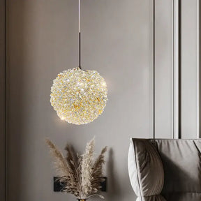 A modern living room features the Morsale.com El Sol Crystal Pendant Light, a spherical chandelier crafted from premium materials that emits a warm, elegant illumination. Below, an arrangement of dried pampas grass in a vase is situated next to a cushioned grey armchair, all set against a textured grey wall.