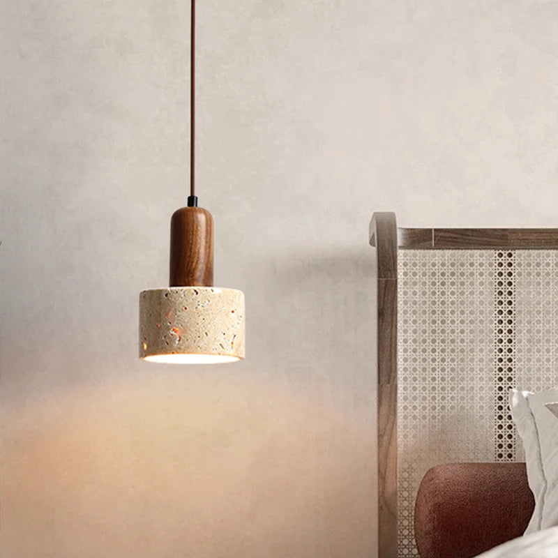 A minimalist kitchen features two Morsale.com Natural Travertine Pendant Lamps with wooden tops and stone-like concrete shades, illuminated by LED bulbs, hanging above a light wood table with a shallow wooden bowl. The background includes a marble backsplash and a counter with minimal décor.