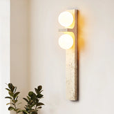 A modern Japan Style wall-mounted light fixture from Morsale.com, featuring two glowing circular LED bulbs, is vertically installed on a light-colored wall. Below the Natural Travertine Modern Wall Lights, to the left, a green leafy plant is partially visible.