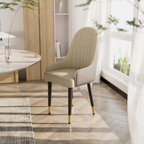 A Morsale Vegan Leather Dining Chair from the set of 2, featuring a modern beige finish, quilted backrest, and black legs with gold tips, rests on a herringbone wooden floor next to a round marble table. Sunlight streams through large windows with sheer curtains, illuminating the space.