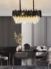 A modern dining room highlights the Morsale Gio Modern Chandelier in black and gold with a crystal shade, elegantly reflecting above the table, which is adorned with a decorative vase of branches, a globe, and books. Gray chairs surround the table, while a paneled wall enhances the contemporary elegance.