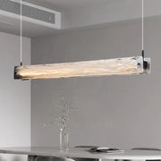 The minimalist room features a Costa Dining Room Linear Suspension Chandelier by Morsale, with a modern luxury design and frosted glass. Below, a dining table holds a slender glass vase with delicate branches. The light gray walls enhance the clean aesthetic provided by energy-saving LED bulbs.