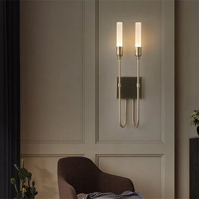 A modern wall showcases the Morsale Candela Contemporary Wall Sconce, featuring cylindrical glass shades that cast a warm ambiance. Beneath the fixture, a brown upholstered chair sits invitingly. A curtain and small plant add subtle touches to this exquisite lighting arrangement in the interior design.