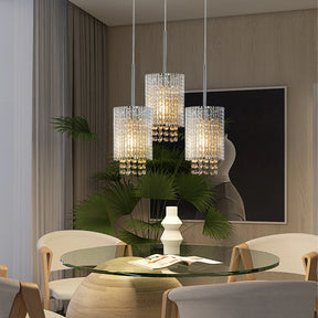 A modern dining room showcases a round glass table with beige chairs. Three Morsale Alleri Crystal Pendant Lights hang overhead, adding elegance. A large potted plant and minimalist black-and-white artwork complete the decor.