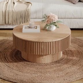 A cozy living room scene features the 31" Round Coffee Table With Storage by Morsale on a woven rug, displaying a vase of pink flowers, a candle, and a book. In the background, a cream sofa with a fringed throw enhances the inviting atmosphere.
