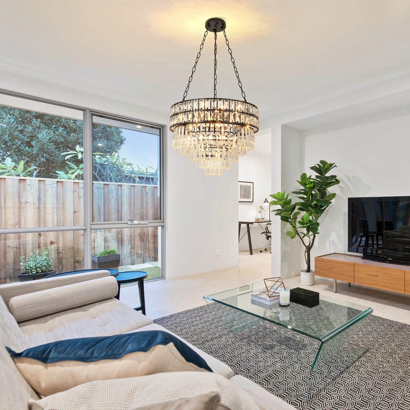 A modern living room features a glass coffee table on a gray rug, a large sofa with cushions, and a flat-screen TV on a wooden stand. Pendant lights complement the Gio Modern Crystal Chandelier by Morsale above, while a window reveals the garden view next to the large potted plant.