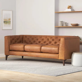The Amalfi 89" Full Grain Leather Couch by Morsale, in a tan tufted design, sits elegantly on a light gray rug. A white wall with minimalist shelves holds books and decor items in the background, complemented by a small white ottoman completing the charming setting.