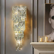 The Bacci Crystal Contemporary Wall Sconce by Morsale is mounted on a gray paneled wall, featuring cascading prisms and illuminated by LED bulbs, casting a warm glow. To the right is part of a black and white portrait. Below, a white mantel adorned with gold candle holders enhances the modern style ambiance.