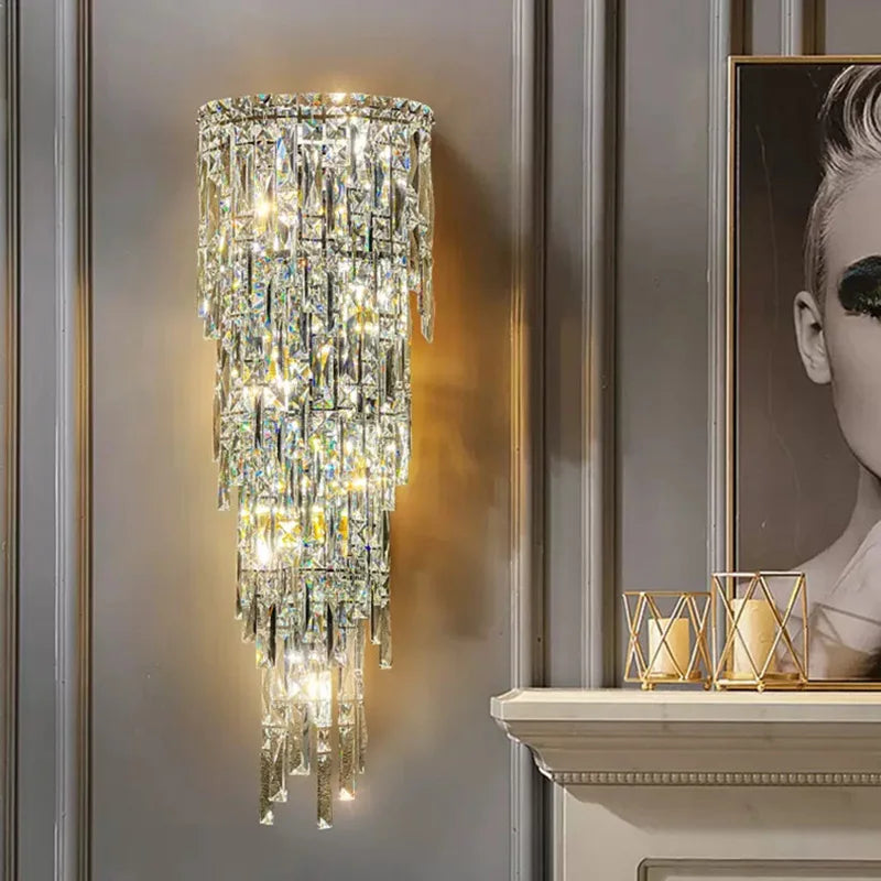 The Bacci Crystal Contemporary Wall Sconce by Morsale is mounted on a gray paneled wall, featuring cascading prisms and illuminated by LED bulbs, casting a warm glow. To the right is part of a black and white portrait. Below, a white mantel adorned with gold candle holders enhances the modern style ambiance.