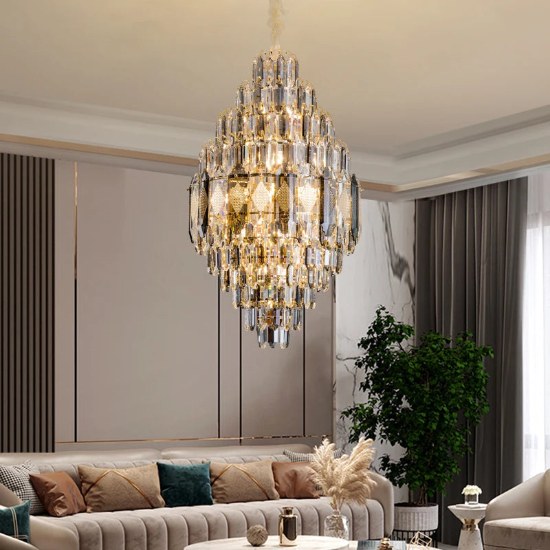 A luxurious living room is highlighted by a large Lazzo Crystal Modern Chandelier from Morsale, with LED bulbs providing a warm glow from above. This modern setup also includes a beige sectional sofa, a round coffee table, and a green potted plant positioned by the window, which is dressed with light gray curtains.