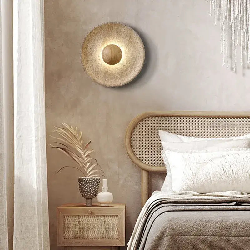 A cozy bedroom with beige walls, a rattan headboard, neutral bedding, and a wooden nightstand featuring a vase with dried plants, illuminated by the Morsale Oakley Natural Stone & Wood Wall Light casting a soft circular glow.