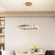 A modern dining room includes a round table with four chairs under a Morsale Natural Travertine Modern Light Fixture. A vase of flowers and bowl of lemons adorn the table. Built-in shelves filled with books line the left wall, enhancing the cozy yet stylish ambiance.