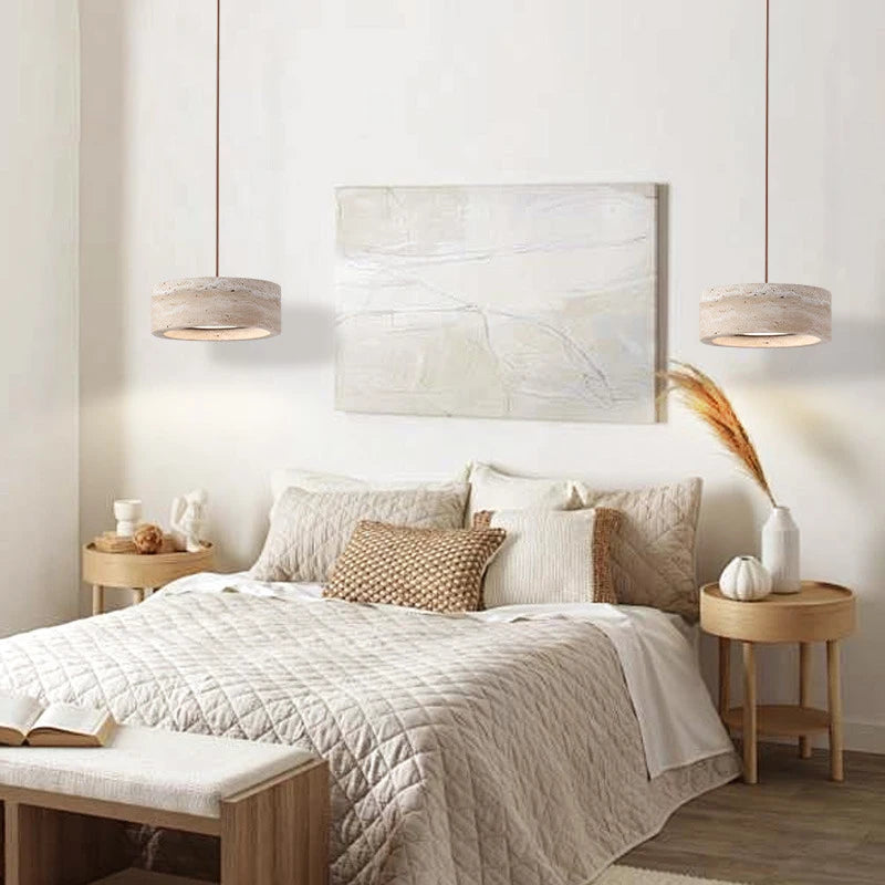 In a minimalist bedroom, a cozy bed with beige pillows and quilt is complemented by two round wooden side tables displaying decor items. Morsale's Modern Travertine Pendant Lights illuminate the room, while an abstract painting adds to the serene ambiance.