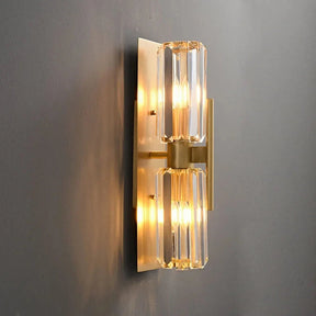 The Morsale Gio Double Head Crystal Wall Sconce showcases a modern design with brass accents and two cylindrical crystal shades. When illuminated, it casts a warm glow against a gray wall, perfect for enhancing any contemporary space.