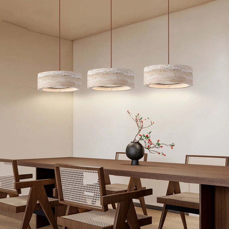 A modern dining area features a wooden table and four chairs with woven backs, illuminated by Morsale's Modern Travertine Pendant Lights. A small black vase with branches and small flowers decorates the table, enhancing the room's minimalist design.