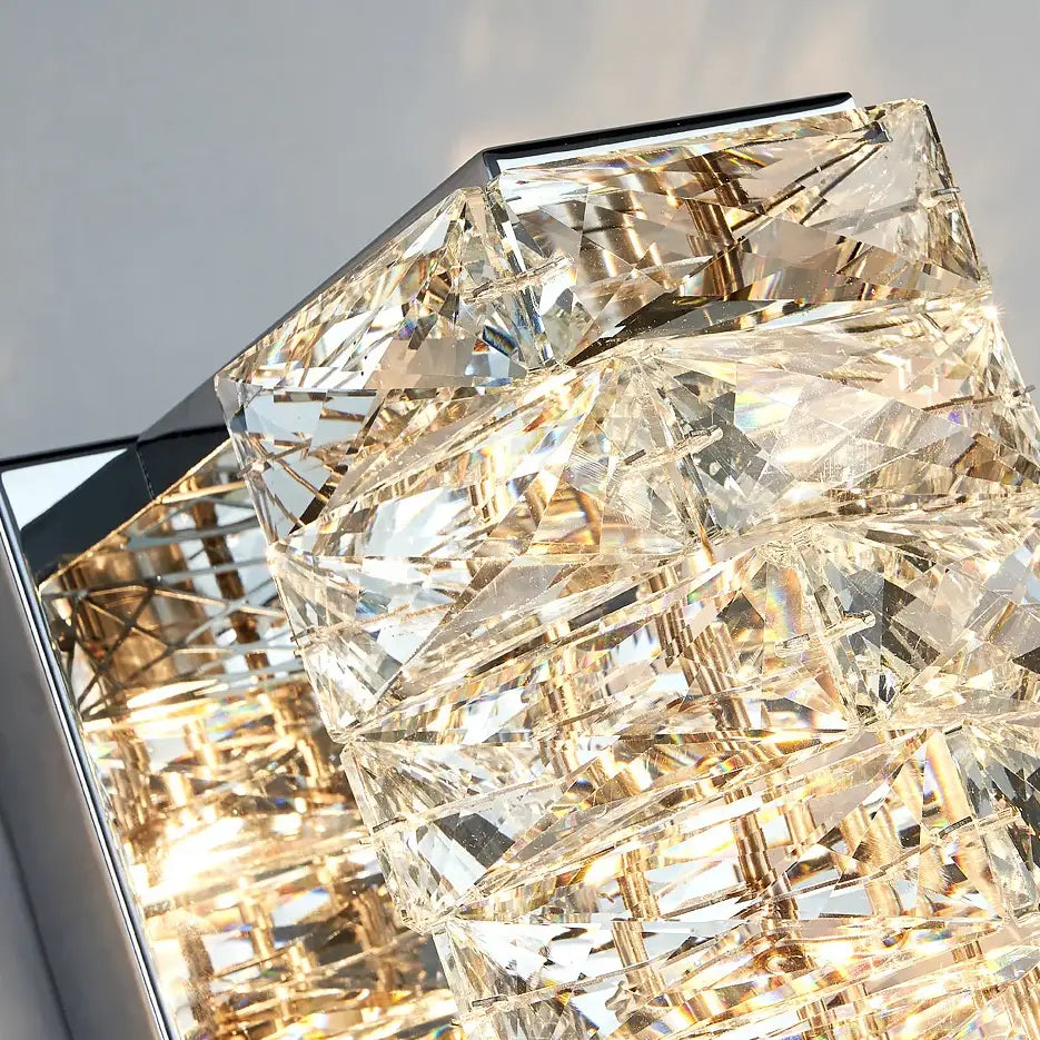 Close-up of the Bacci Crystal Wall Sconces by Morsale.com, featuring reflective, geometric crystal shapes set in a brass frame. Bright, warm light illuminates through and around the crystals, creating a prismatic effect on the surrounding surfaces and exemplifying elegant modern design.
