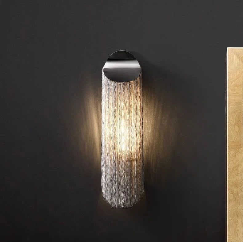 A modern Morsale.com Cadena Chain Tassel Wall Sconce, featuring a black mount and cascading metallic chains, illuminates against a dark wall. Below it sits a beige couch adorned with multiple cushions, positioned next to a light wooden panel on the right. This sleek aluminum design adds sophistication to any living space.