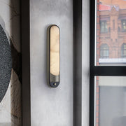 The Morsale Sandford Stainless Steel & Marble Wall Sconce elegantly complements a textured gray wall, casting a soft, warm glow. Next to a window, it frames an urban view of red brick buildings with large windows in the background.