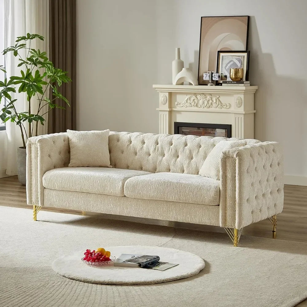 A modern living room features a Morsale Button Tufted Sofa Couch in cream composite fabric with gold legs, paired with a matching pillow and round rug. A decorative fireplace artfully adorned with vases sits in the background, and a lush green plant enhances the window area.
