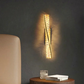 The Morsale Bacci Crystal Wall Sconce, featuring a twisted design, emits a soft glow on a dark wall. Beneath it, a round wooden table displays a clear glass and a closed book, while part of a light-colored chair can be seen to the left. The LED bulbs provide energy-efficient illumination.