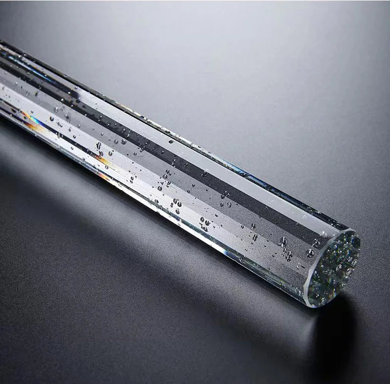 A clear acrylic or glass rod lies diagonally on a dark grey surface. The rod, resembling an elegant Modern Minimalist LED Pendant Light by Morsale.com, contains small air bubbles suspended within it, adding texture and visual interest. The surface of the rod reflects light like polished gold, creating subtle highlights.