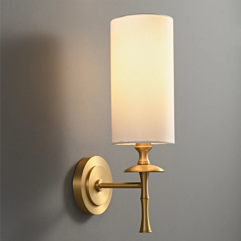 The Morsale American Retro Copper Wall Light, with its cylindrical white fabric shade, is elegantly mounted on a gray wall. Featuring LED lighting, it emits a soft and warm glow, offering a modern and sophisticated design.