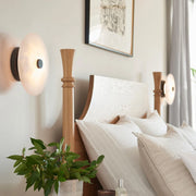 A neatly made bed with white bedding and multiple pillows is flanked by two Morsale Witney Copper & Marble Wall Sconces. A wooden headboard and framed picture above enhance the decor, while a vase with green leaves on a bedside table completes the cozy, chic ambiance.
