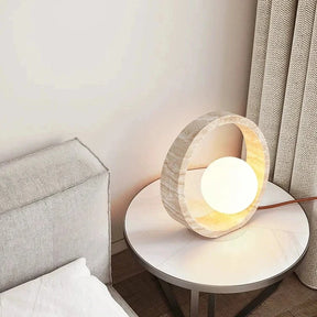 The Modern Travertine LED Table Lamp by Morsale.com features a round Natural Yellow Cave Stone frame with a textured, natural finish and a glowing spherical light bulb in the center. The lamp emits a warm, ambient light, creating a cozy atmosphere, and has a cord extending from the back.