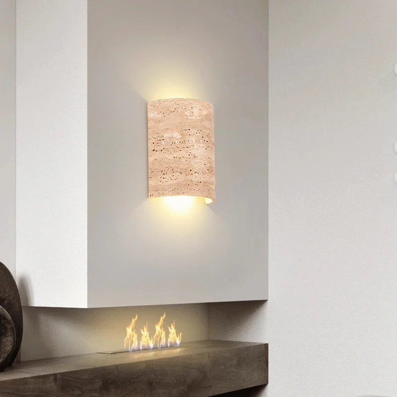 A Natural Travertine Art Deco Wall Lamp from Morsale.com, crafted from beige, porous stone and shaped into a cylinder, is mounted on a white wall. The light radiates softly from both the top and bottom, generating an ambient glow around the fixture that accentuates its ART DECO design.