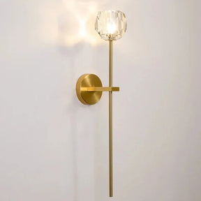 The Clear Crystal & Brass Wall Light Fixture by Morsale.com features an elegant modern design with a gold cylindrical base and a long, sleek gold rod extending vertically. At the top of the rod is a clear crystal and brass lampshade that emits warm, soft ambient lighting against a plain white wall.