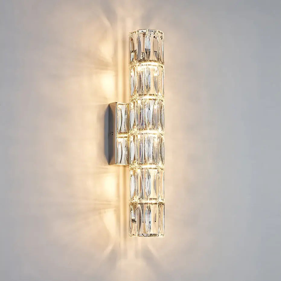 The Bacci Crystal Wall Light Sconce by Morsale.com is a vertical wall sconce with a contemporary design, featuring a row of handmade crystal blocks that evenly diffuse the warm light emitted from behind them. This elegant indoor lighting fixture is mounted on a metallic base and attached to a neutral-colored wall.