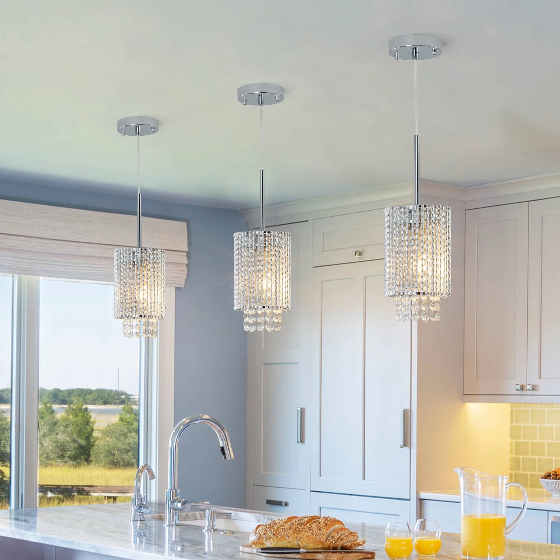 Above the marble island, Morsale's Alleri Crystal Pendant Lights add modern elegance with easy installation. The island holds baked goods and a pitcher of orange juice, while the window reveals a lush landscape outside.