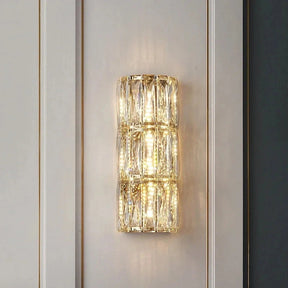 A Bacci Crystal Wall Light from Morsale.com, featuring three vertically aligned rectangular sections, casts a warm and diffused light against a white paneled wall. Below, a green velvet sofa adorned with white cushions and accompanied by a glass side table holding a potted plant and decorative items enhances the elegant ambiance.