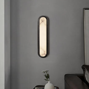 A Moonshade Natural Marble LED Wall Lamp by Morsale, featuring a modern vertical design with a sleek black frame and a white illuminated center, hangs on a grey wall. Below it, a small side table holds a white vase with a few delicate flowers. Part of a black sofa is visible in the bottom right corner.