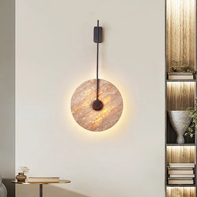 A contemporary wall showcases the Moonshade Natural Marble Sconce by Morsale, a circular marble lamp featuring a golden, textured surface that emits a soft glow. This elegant sconce is mounted on a sleek black vertical fixture. Adjacent are a small round table, an ornamental vase, and a bookshelf embellished with books and plants.