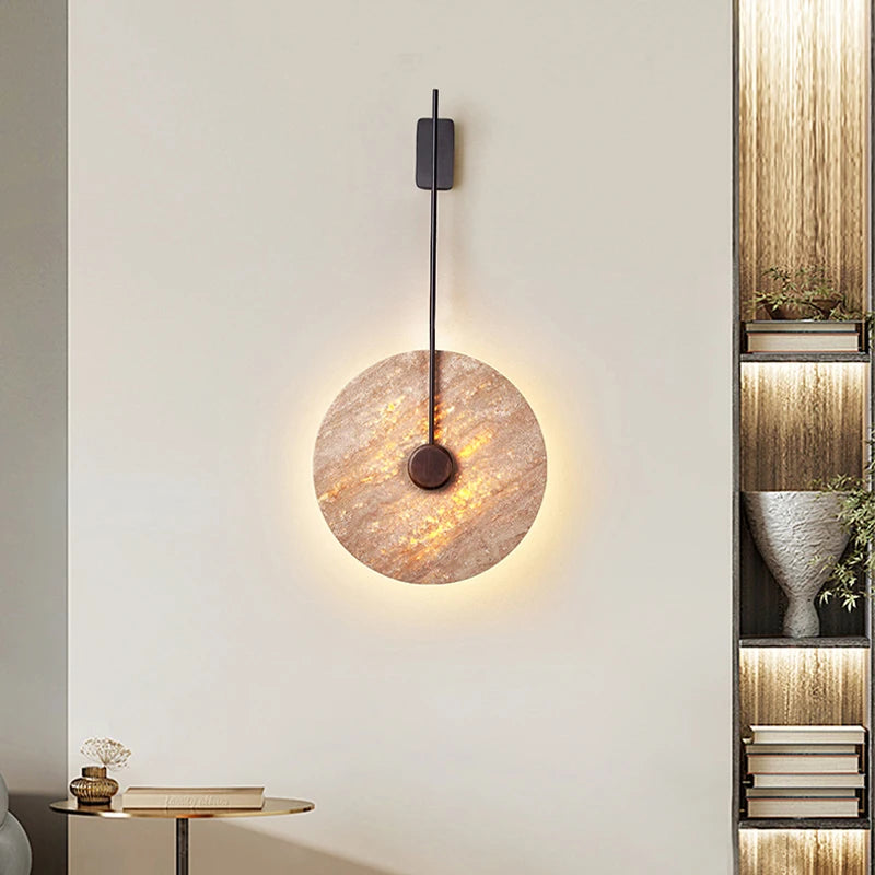 A contemporary wall showcases the Moonshade Natural Marble Sconce by Morsale, a circular marble lamp featuring a golden, textured surface that emits a soft glow. This elegant sconce is mounted on a sleek black vertical fixture. Adjacent are a small round table, an ornamental vase, and a bookshelf embellished with books and plants.