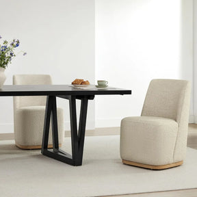 A modern dining area showcases a dark rectangular table, two Morsale Boucle Dining Chairs With Casters in off-white, on a light carpet. A vase of flowers and a plate of pastries accompany a green cup, against white walls and light wood flooring.
