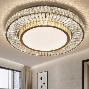 The Morsale Gio Crystal Ceiling Chandelier, a modern circular light fixture adorned with layers of crystal-like designs and equipped with LED bulbs, emits a warm glow in a contemporary room. The room features light walls, a window with curtains, and a framed abstract art piece.